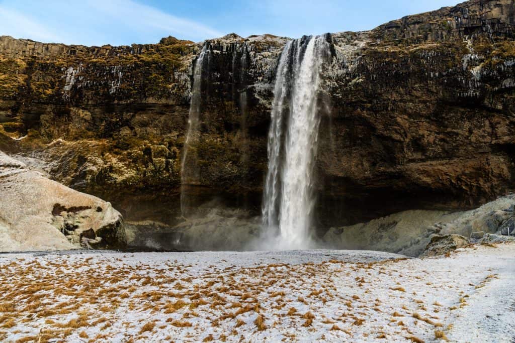 A tall waterfall plunges from a moss-covered cliff into a shallow basin, with patches of frost and snow blanketing the rocky ground below. The cliff face is lined with icicles, adding to the wintry feel of this dramatic Icelandic landscape.
