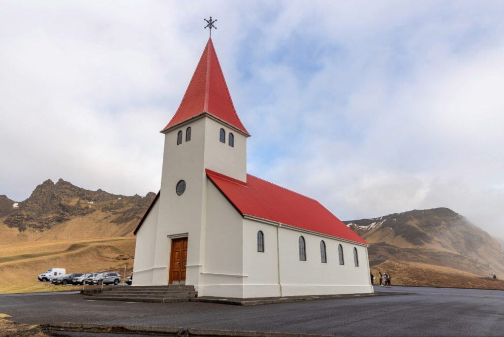 A white church with a steep red roof and central steeple stands against a dramatic mountain backdrop under a partly cloudy sky. The building has arched windows and a wooden front door, with a few parked cars and scattered visitors nearby, emphasizing its remote and scenic location.