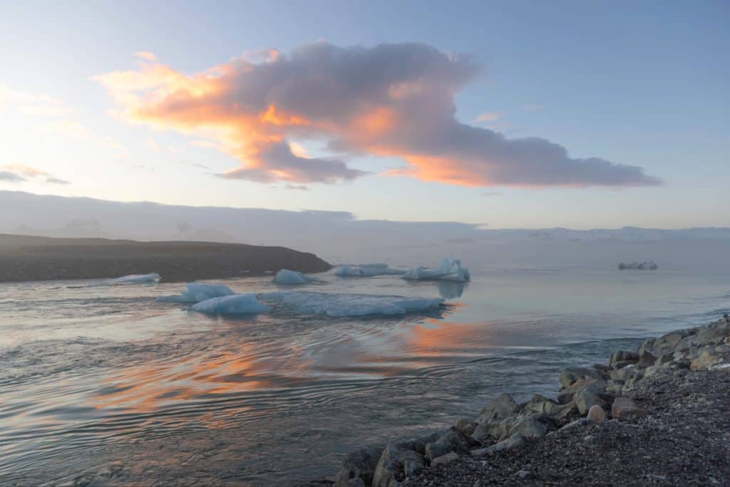 Floating icebergs drift through a calm glacial lagoon at sunset, their blue hues contrasting with the soft pink and orange clouds reflected in the water. Rocky shoreline and distant mountains frame the tranquil Icelandic landscape.