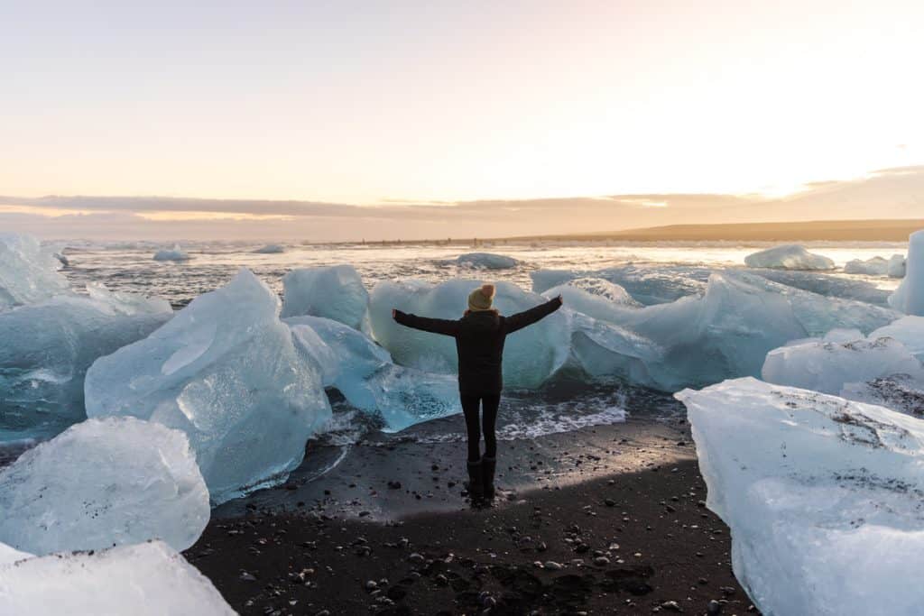 A person in winter clothing stands with arms outstretched on a black sand beach, surrounded by large, glistening icebergs washed ashore. The golden light of sunset reflects off the ice and ocean, creating a magical contrast between the icy blues and warm sky.