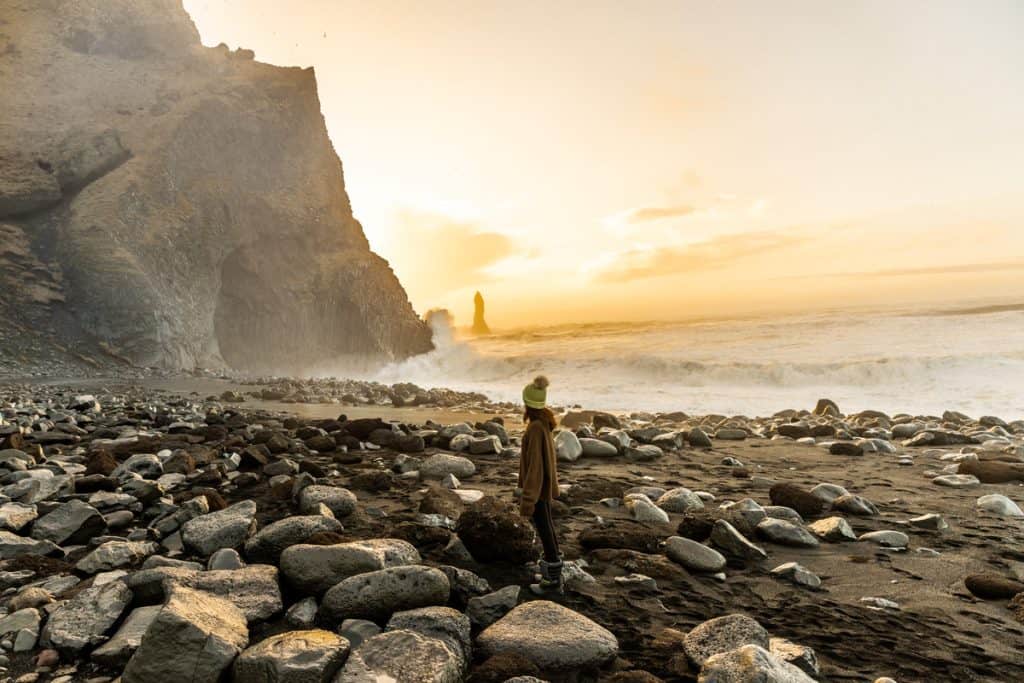 A person in a beanie and boots stands on a rocky black sand beach at sunset, gazing toward crashing waves and a dramatic sea stack rising from the ocean. Golden light bathes the cliffs and shoreline, casting a warm glow across the rugged Icelandic coast.