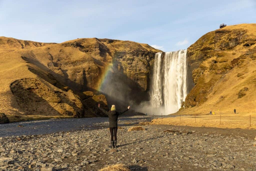 A person in a black coat and beanie stands with arms outstretched facing Skógafoss waterfall in Iceland, with a faint rainbow visible in the mist. The surrounding landscape is rugged and golden-brown, with rocky terrain and steep cliffs under a clear sky.