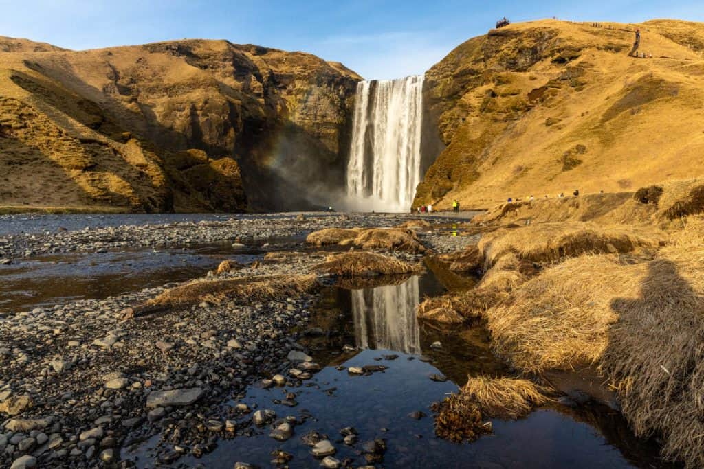 A powerful waterfall cascades down a rugged cliff into a shallow, rocky riverbed, with golden grass and a clear reflection in the foreground. Tourists are visible along the trail and near the base of the falls, emphasizing the scale and natural beauty of the landscape.