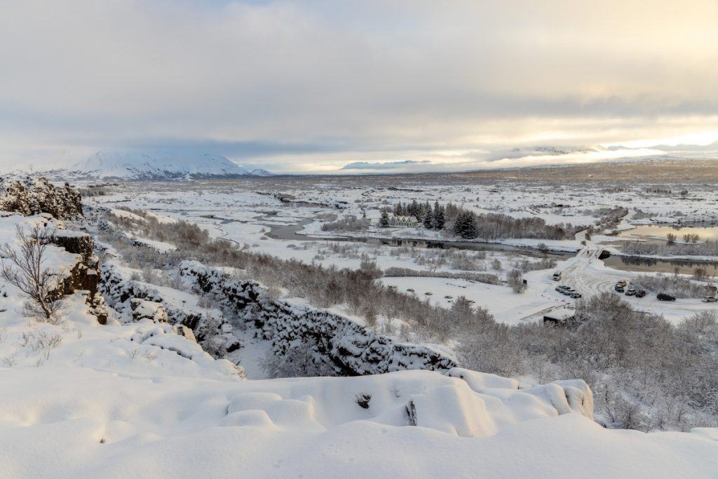 A wide snowy landscape stretches across a valley with winding rivers cutting through the white terrain and mountains rising in the distance. Snow covered cliffs frame the foreground while a small road and parked cars sit near a bridge under a soft winter sunset sky.