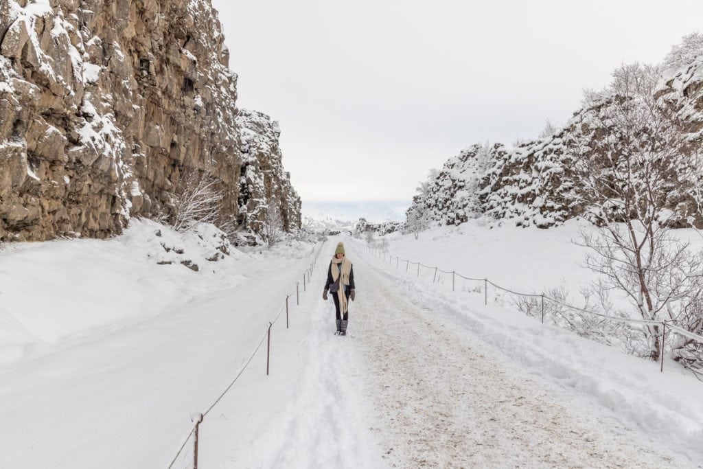 A person bundled in a long coat scarf and knit hat walks down a snow covered path between towering rocky cliffs. The ground and surrounding landscape are blanketed in fresh white snow creating a quiet winter scene with steep canyon walls on both sides.