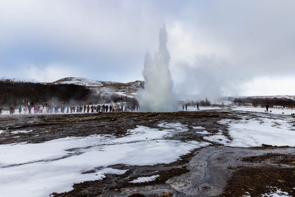 A geyser erupts high into the cold air as a crowd of bundled visitors stands in a semicircle watching from a snowy geothermal field. Steam rises around the group and the dark rocky ground contrasts with patches of white snow in the winter landscape.