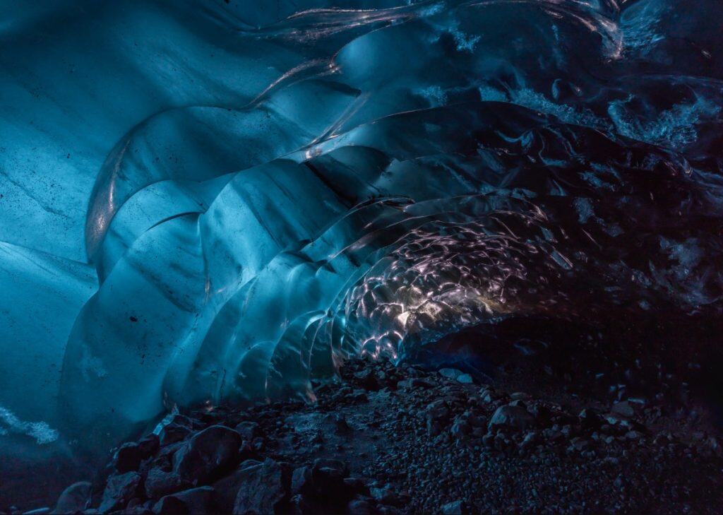Inside a glacial ice cave, soft light reflects off the smooth, rippled blue walls, casting shimmering patterns across the tunnel-like ceiling. The rocky floor adds contrast to the surreal, almost translucent beauty of the frozen interior.