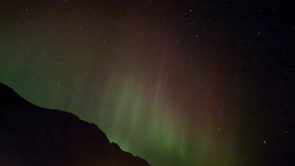 Faint green and reddish aurora lights stretch upward from behind a silhouetted mountain, blending into a star-speckled night sky.