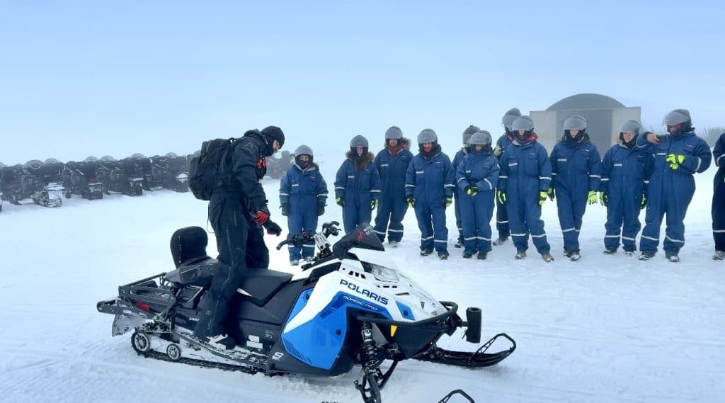 A snowmobile guide dressed in black demonstrates how to operate a Polaris snowmobile while a group of people in matching blue insulated suits and helmets watches in a snowy, foggy landscape. Snowmobiling across glaciers is one of the most thrilling things to do in Iceland.