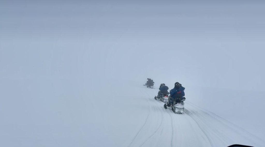 A group of people ride snowmobiles in single file across a vast, snow-covered landscape under a thick layer of fog or whiteout conditions. The scene is minimal and monochromatic, emphasizing the isolation and adventure of traveling through a frozen wilderness.