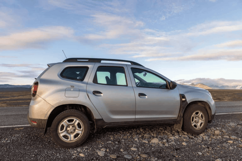 A silver SUV is parked on a gravel shoulder beside an open road with mountains and a soft sunset sky in the background. The vehicle sits in a wide rural landscape suggesting a road trip through a remote scenic area.