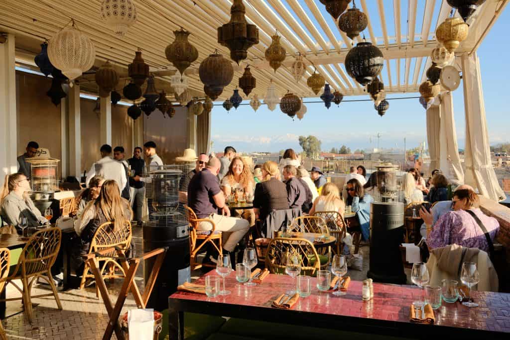 Bustling Marrakech rooftop restaurant at golden hour, with dozens of ornate Moroccan lanterns hanging from a slatted pergola, rattan chairs around small tables filled with diners, and a distant view of the Atlas Mountains on the horizon.