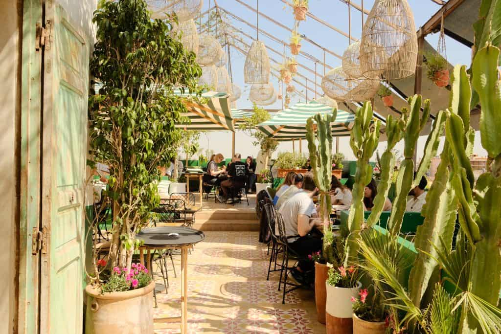 Marrakech rooftop café with green-and-white striped parasols, woven rattan pendant lights, cacti and potted plants framing the space, and patterned encaustic floor tiles, with diners seated at small tables along a sunlit terrace.