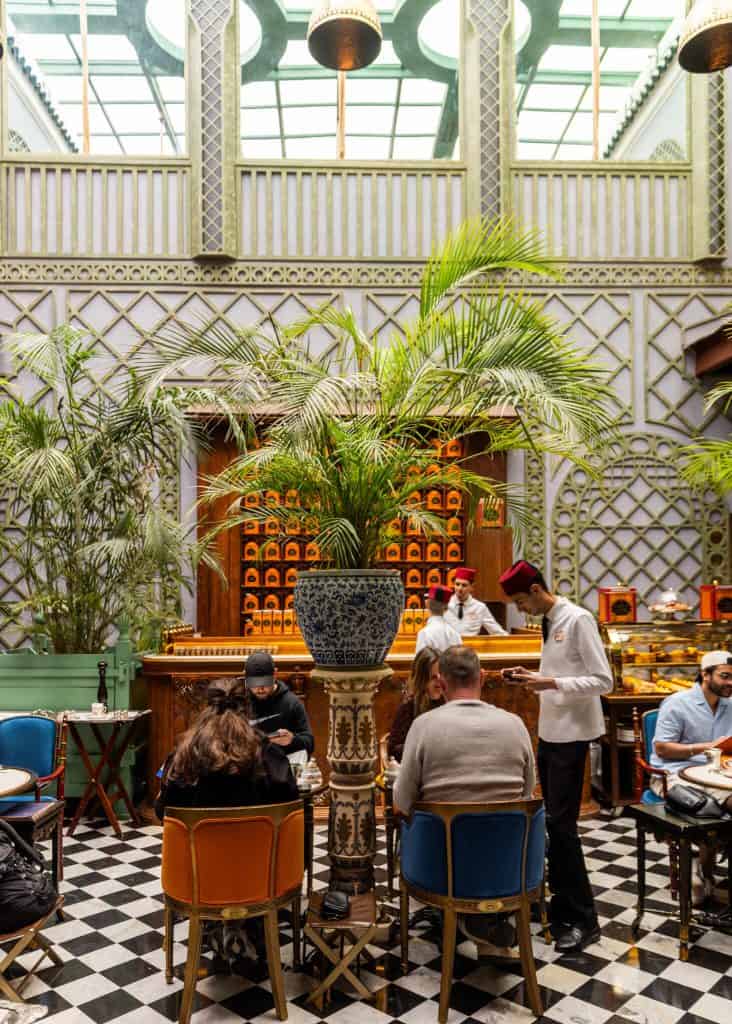 Alt text: Bright indoor café with a glass ceiling and ornate patterned walls, where a large potted palm sits in the center of a tiled floor surrounded by seated guests. A server in a white uniform and red hat takes an order while pastries and tea tins line the wooden counter behind him.