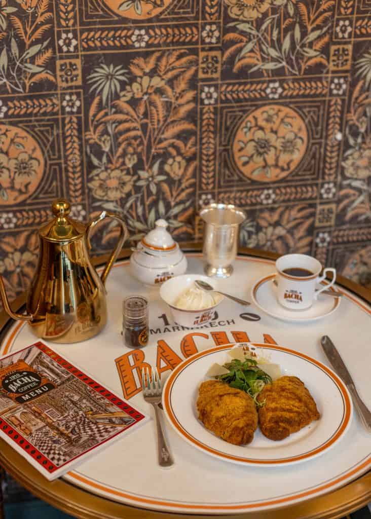 Table setting at Bacha Coffee with a gold teapot, cup of black coffee, and two croissants on a white plate garnished with greens and cheese. A small bowl of whipped cream, a spice jar, and a menu labeled “Bacha Coffee Menu” sit on a tray printed with “Marrakech Bacha Coffee,” against a detailed floral tile wall.