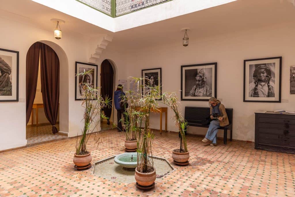 Interior courtyard of a Marrakech riad gallery with a small green-tiled fountain, terracotta floor tiles, potted papyrus plants, and large black-and-white portrait photographs displayed on the white walls. Two visitors browse the exhibition, and an open skylight reveals an ornate wrought-iron balcony above