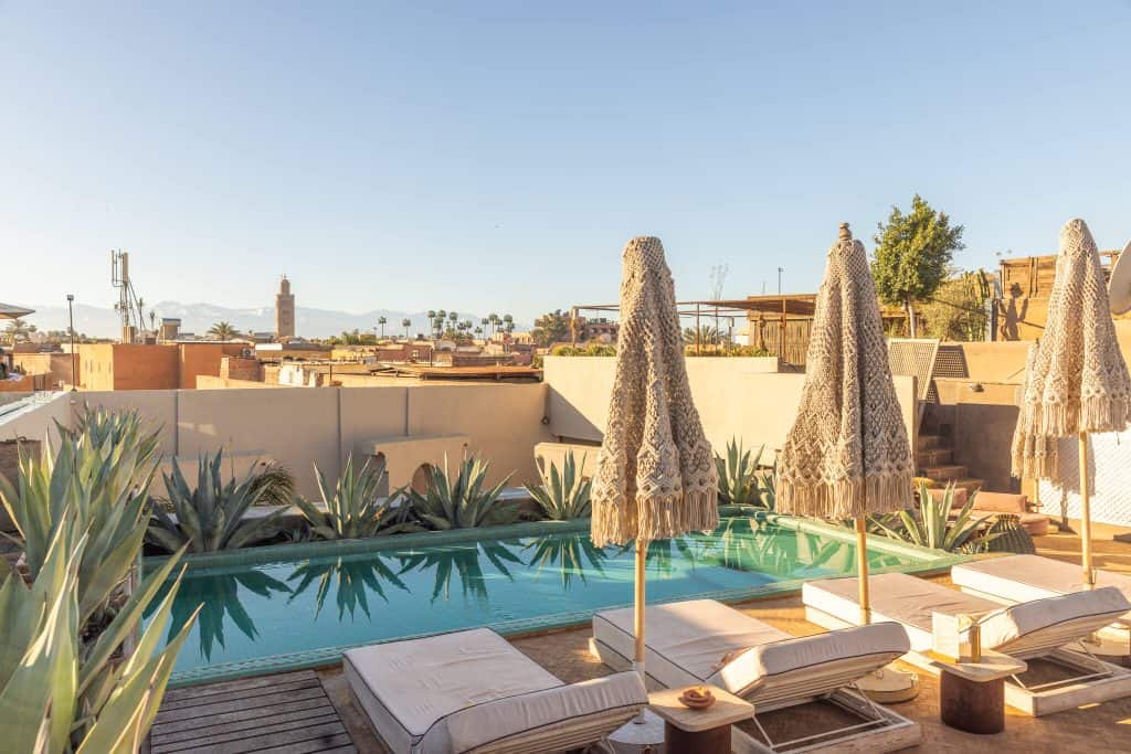 Rooftop terrace with a small rectangular pool surrounded by lounge chairs and tall woven umbrellas, overlooking a sunlit cityscape with palm trees and distant mountains. Neutral-toned walls and desert plants frame the space, creating a calm setting for relaxing above the bustling streets.