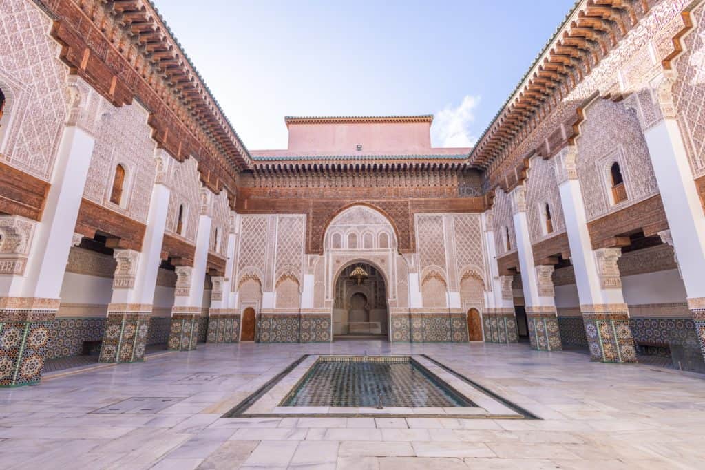 The grand courtyard of the Ben Youssef Madrasa in Marrakech, with a long rectangular reflecting pool at its center, marble-paved floors, and walls covered in elaborately carved stucco, cedar woodwork, and geometric zellige tilework. A horseshoe-arched prayer hall entrance sits at the far end beneath a clear blue sky.