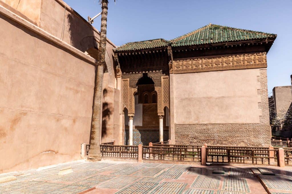 Moroccan mausoleum entrance with carved stucco, marble columns, and a green-tiled roof, set into a terracotta wall above a courtyard of tiled tomb markers.