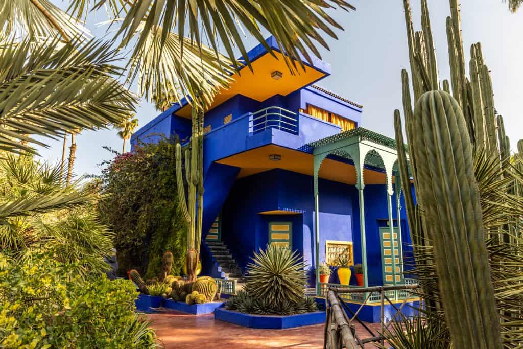 The cobalt blue Art Deco villa at Jardin Majorelle in Marrakech, with bright yellow window frames, a green-arched veranda, and an exterior staircase, surrounded by towering cacti, agaves, and palm trees.