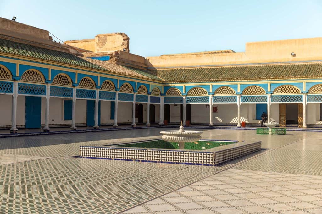 Sunlit Moroccan palace courtyard with a white tiered fountain, checkered tile basin, geometric floor tiles, and a turquoise-and-yellow arcaded gallery with green-tiled roof.