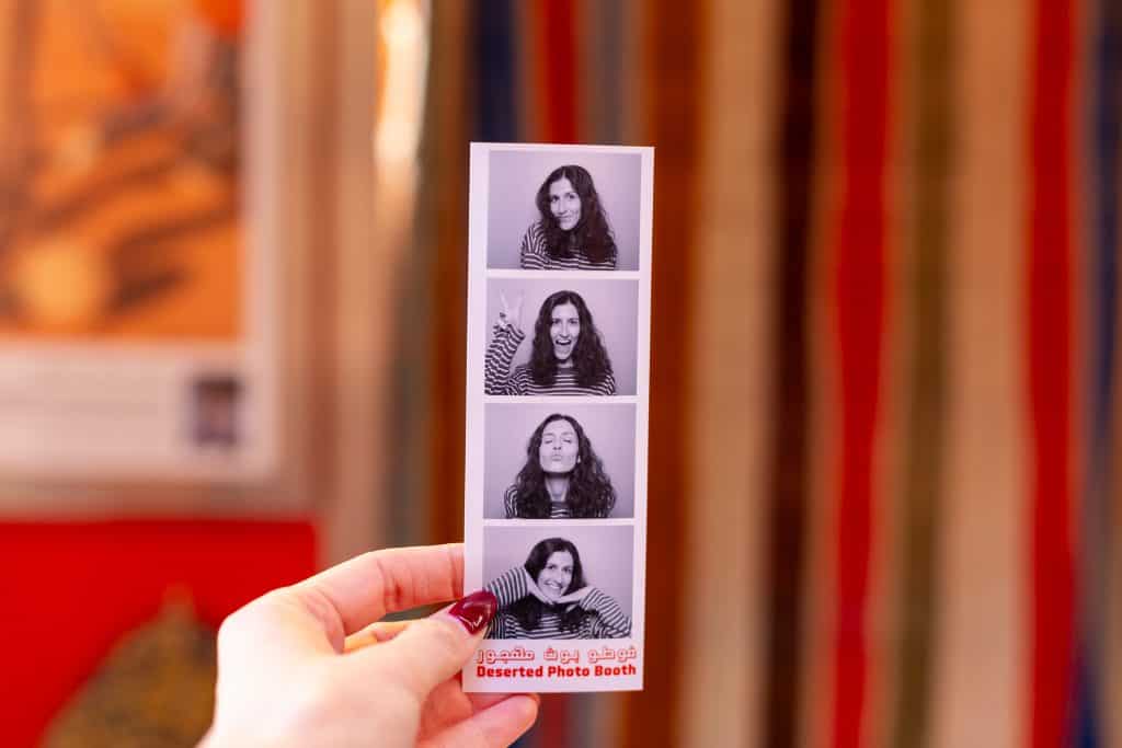 A hand with dark red nail polish holds up a black-and-white photo booth strip showing four playful portraits of a woman with long dark hair in a striped top, captioned 'Deserted Photo Booth' in red with Arabic script above. The background is softly blurred in warm reds and golds.