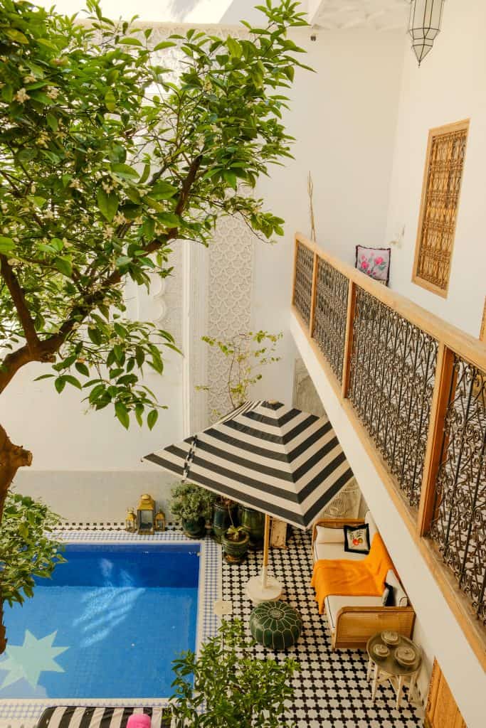 Overhead view of a Marrakech riad courtyard with a blue-tiled pool featuring a star motif, a black-and-white striped umbrella shading a rattan sofa with an orange throw, black-and-white patterned floor tiles, and a blossoming citrus tree reaching up past a wrought-iron balcony.