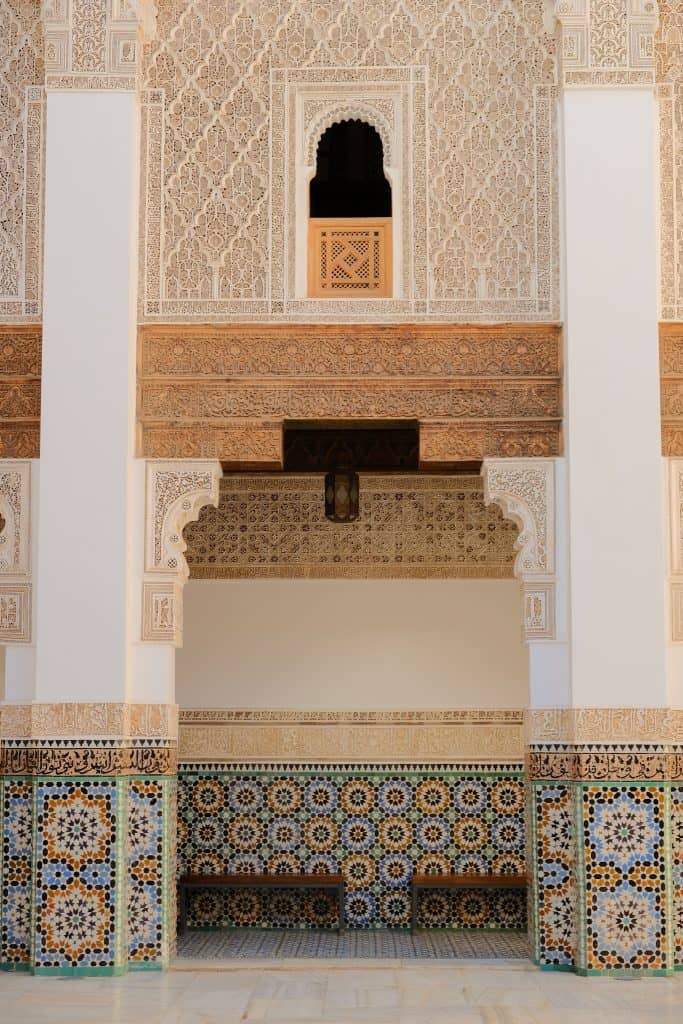 Interior wall of the Ben Youssef Madrasa in Marrakech, layered with intricately carved stucco, Arabic calligraphy bands, a small latticed wooden window, and a base of vibrant geometric zellige tilework in blue, green, and ochre.