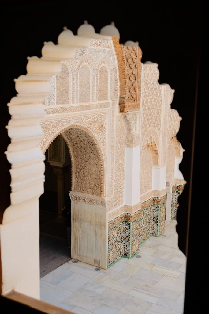 View through a scalloped horseshoe arch into the courtyard of the Ben Youssef Madrasa, revealing finely carved stucco walls, a star-patterned zellige tile dado, and a marble-paved floor.