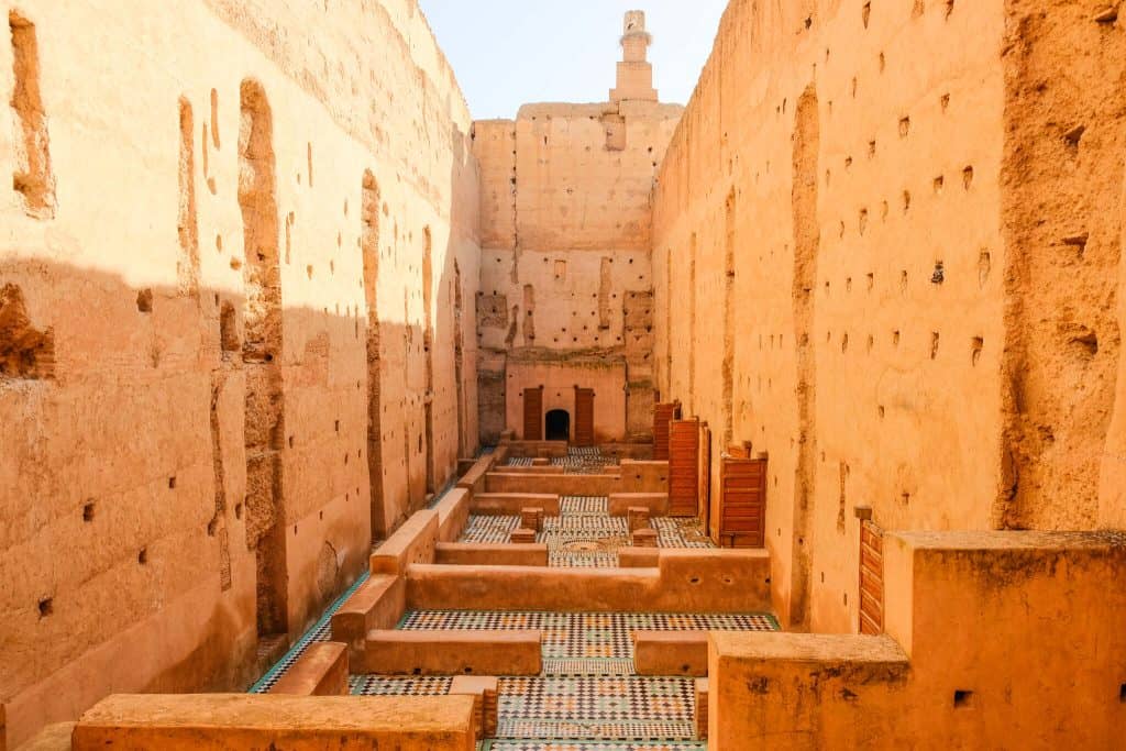 Ruins of El Badi Palace in Marrakech, with towering weathered ochre rammed-earth walls flanking a sunken chamber lined with patterned mosaic tile floors, and a stork's nest perched atop a distant tower.