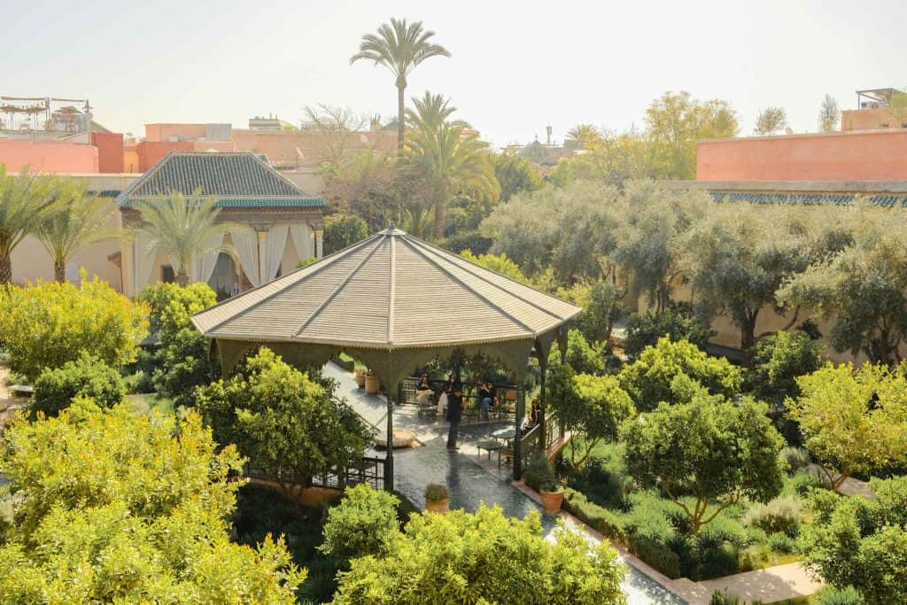 Elevated view of a sunlit Marrakech garden courtyard with an octagonal pavilion at its center, surrounded by lush citrus trees, palms, and terracotta walls, with a few visitors seated inside the pavilion