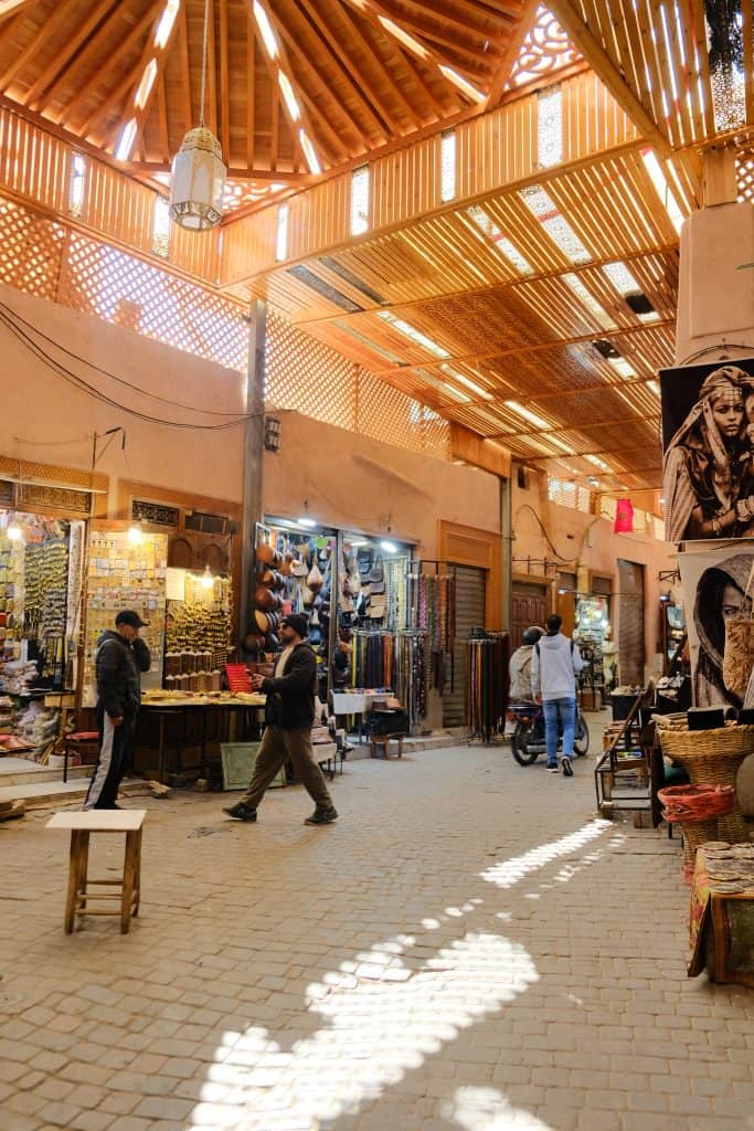Narrow cobblestone alley in the Marrakech medina lined with shops displaying colorful woven handbags and baskets, with shoppers and a cyclist passing under a slatted wooden roof as sunlight streams through.