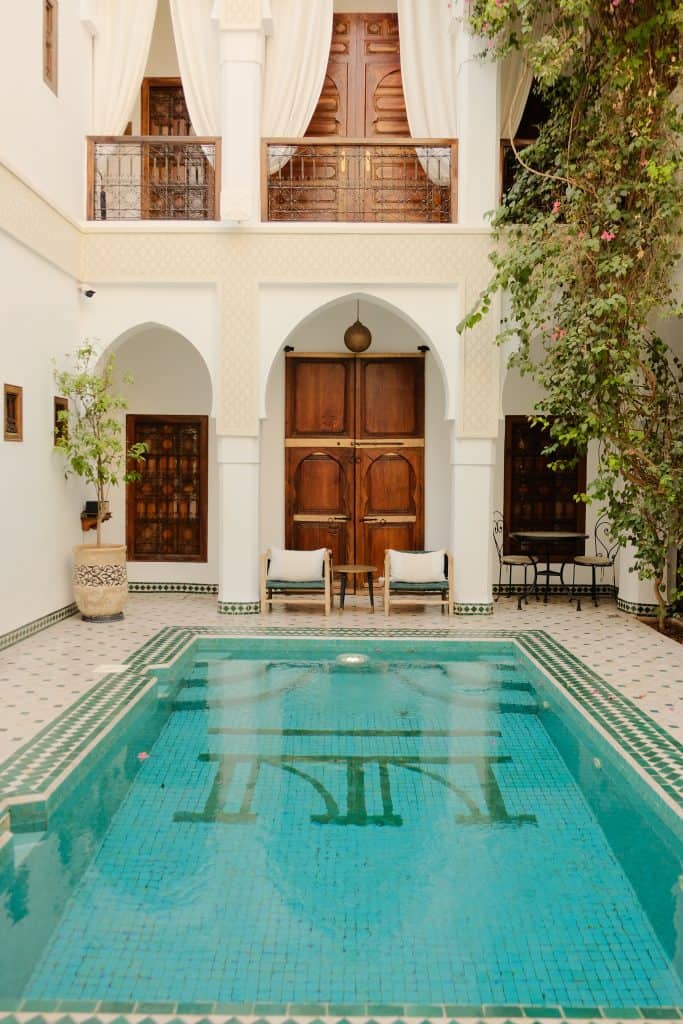 Riad courtyard in Marrakech with a small turquoise-tiled plunge pool, framed by white horseshoe arches, carved wooden doors, sheer curtains, and a flowering bougainvillea climbing one wall.