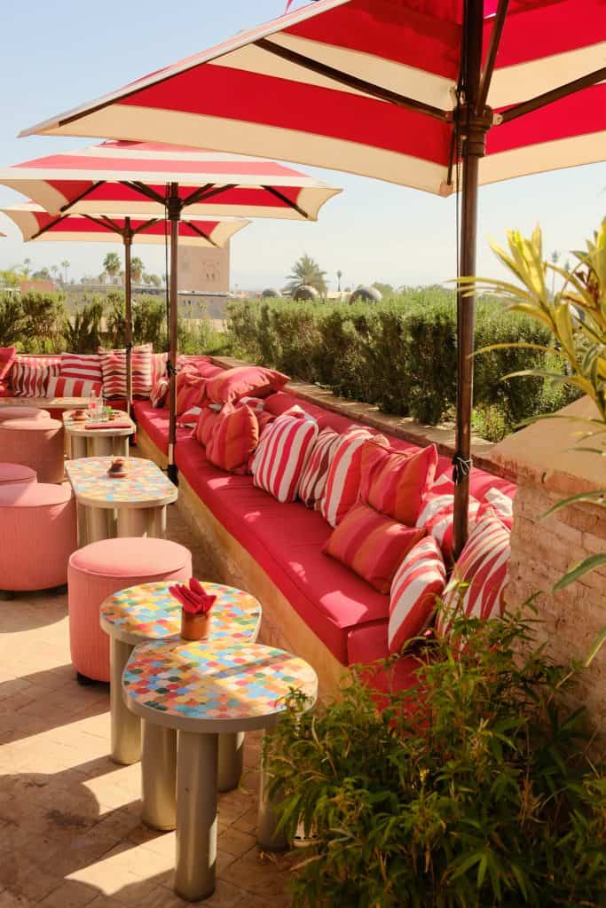 Image 1: "Marrakech rooftop terrace with red-and-white striped umbrellas shading a long banquette piled with pink and striped cushions, colorful mosaic-tiled side tables, and a hedge-lined view toward the Koutoubia Mosque in the distance.
