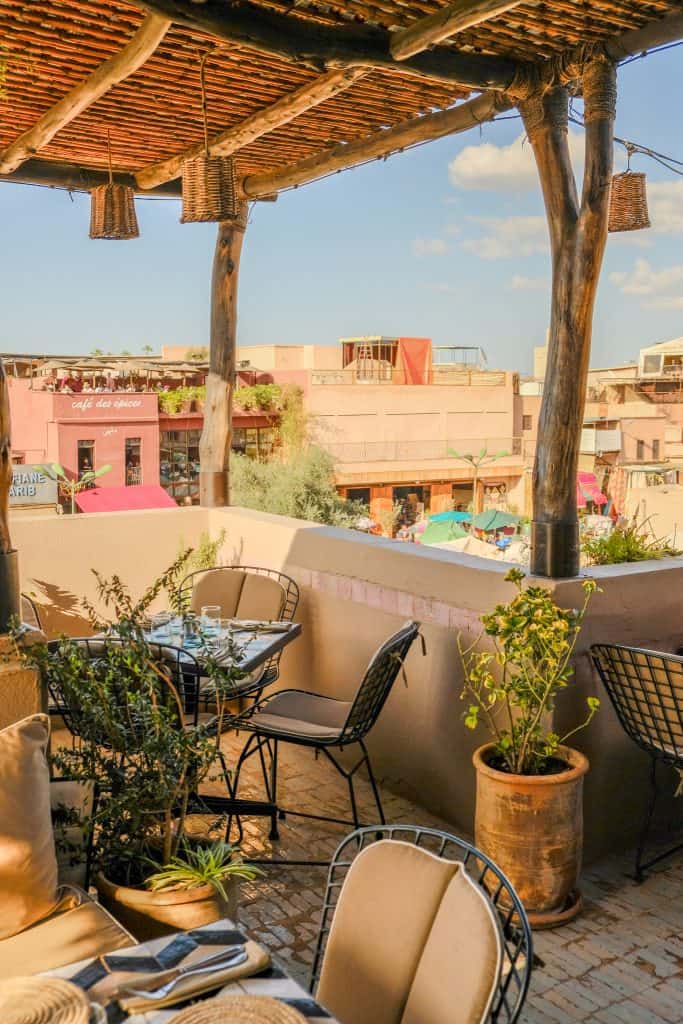 Marrakech rooftop café with a rustic reed-and-wood pergola, woven hanging lantern baskets, black wire chairs at small dining tables, and a view across to Café des Épices and the bustling square below.