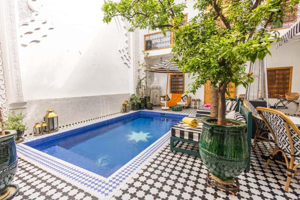 Classic riad courtyard in Marrakech with a star-tiled plunge pool, black and white geometric floor tiles, orange trees and Moroccan lanterns.