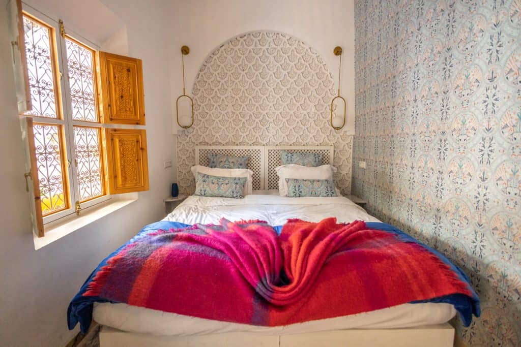 Marrakech riad bedroom with a double bed beneath an arched patterned headboard, brass wall sconces, pastel block-printed wallpaper, and a vibrant pink and blue Berber blanket folded at the foot. Wooden shutters frame an ornate wrought-iron window.
