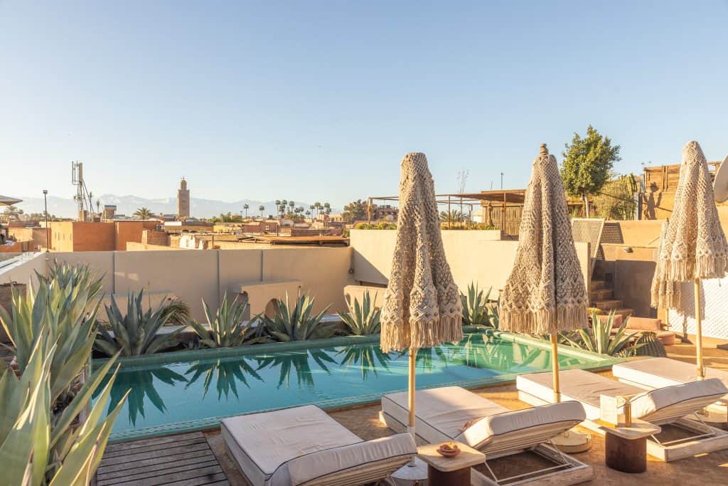 Marrakech riad rooftop with a long turquoise pool lined by agave plants, white sun loungers, and three macramé-fringed parasols, with a view across the medina to the Koutoubia Mosque and the snow-capped Atlas Mountains.