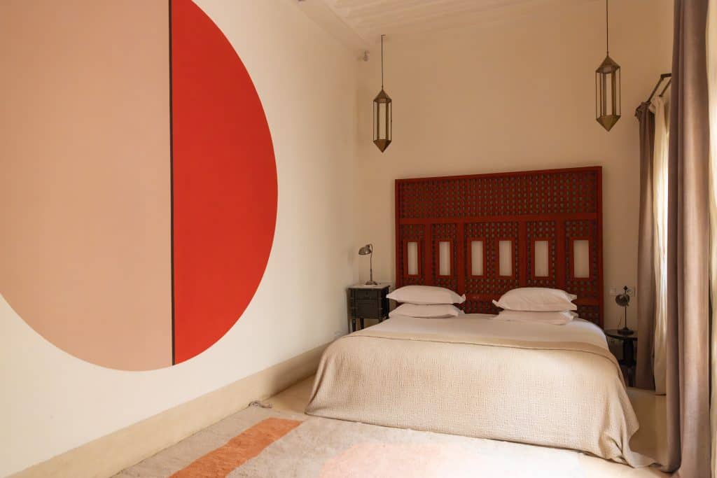 Minimalist Marrakech riad bedroom with a wooden mashrabiya headboard painted red, a large pink-and-red circle mural on the white wall, brass pendant lanterns, and a neutral textured throw on a low bed.