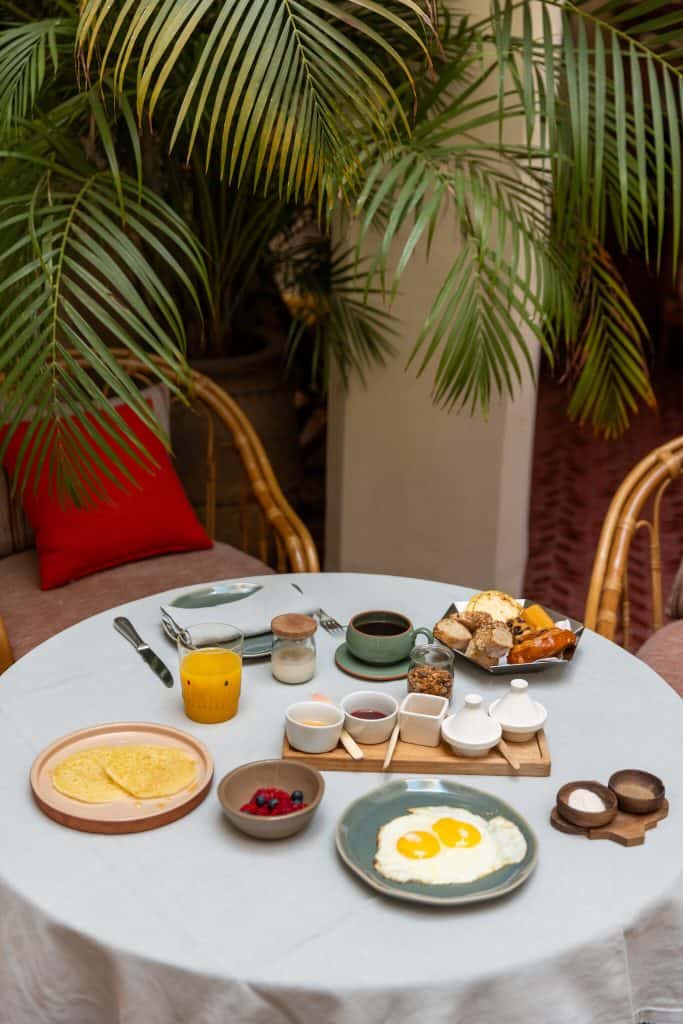 Moroccan breakfast spread on a round table in a riad courtyard, with fried eggs, a crepe, a bowl of berries, fresh orange juice, coffee, and a wooden board of jams and condiments, set among rattan chairs and palm fronds.