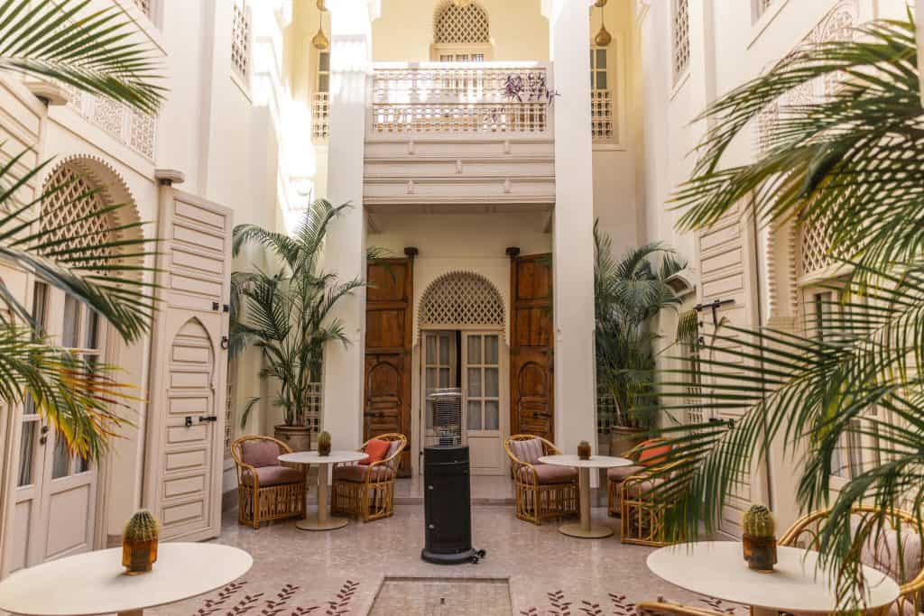 Bright Marrakech riad courtyard with cream walls, intricately carved white shutters, rattan armchairs with pink cushions around small white tables, tall potted palms, and a balcony with a latticed railing above an arched wooden doorway.