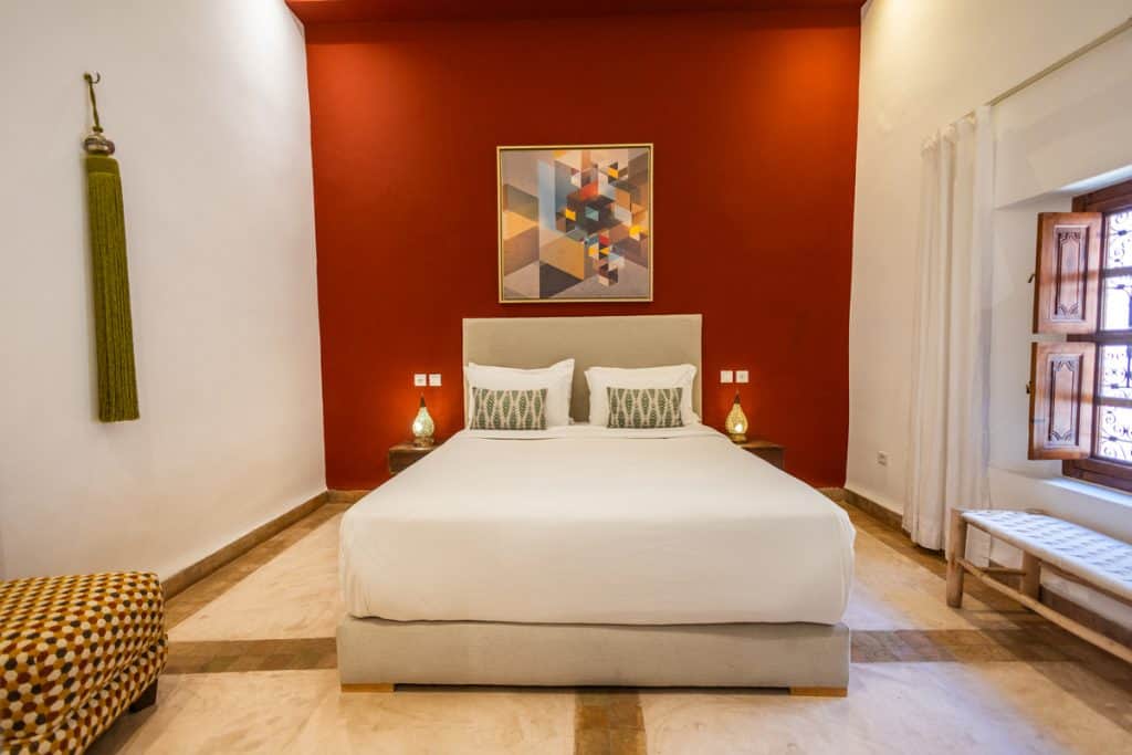 Marrakech riad bedroom with a deep terracotta accent wall behind a cream upholstered bed, a colorful geometric artwork above, gilded teardrop bedside lamps, and a carved wooden window letting in soft light.