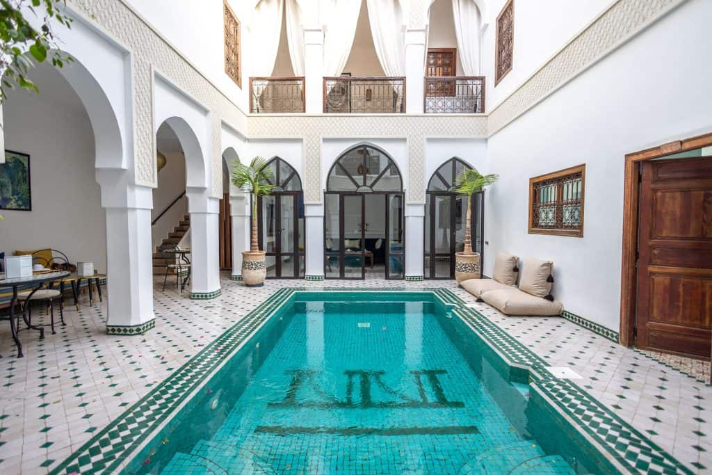 Marrakech riad courtyard with a large turquoise mosaic-tiled pool at its center, flanked by white horseshoe arches, tall arched windows, and potted palms. A wrought-iron balcony with flowing white curtains looks down from above, and cream cushioned seating lines the edge.