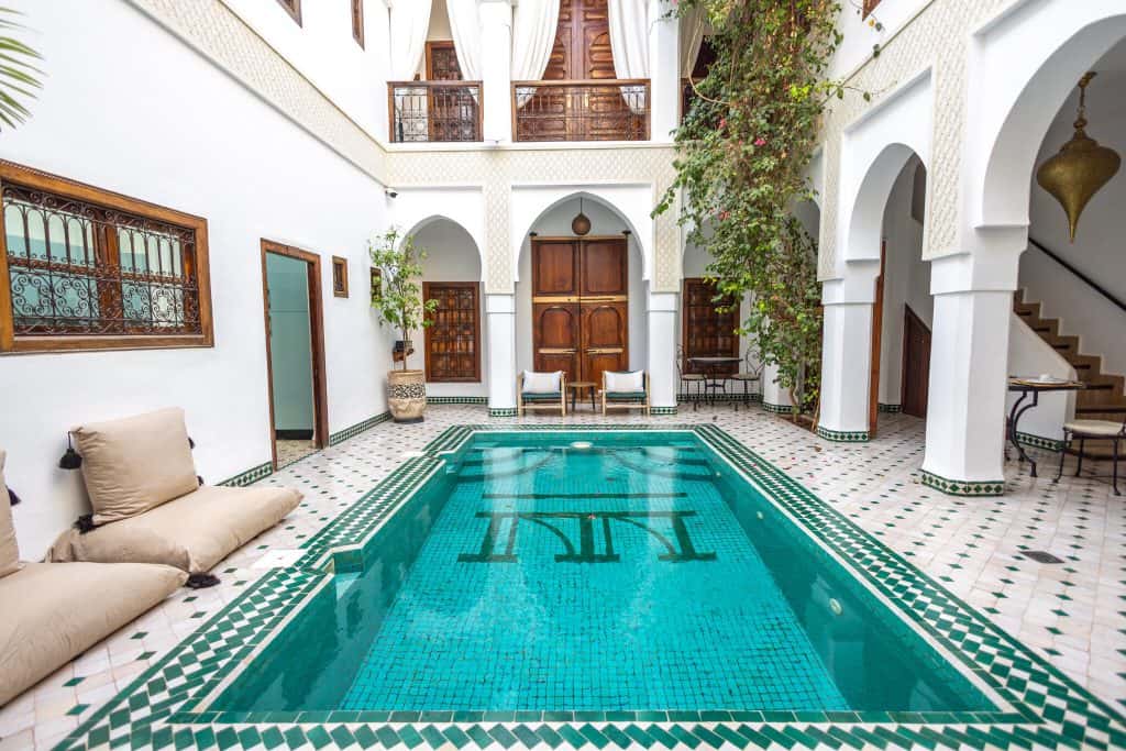 Symmetrical riad courtyard in Marrakech with a turquoise tiled plunge pool, white Moorish arches, carved wooden doors and floor cushions for lounging.