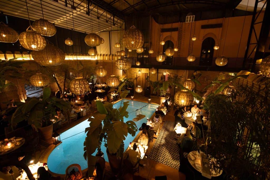 Atmospheric Marrakech restaurant at night, with dozens of ornate golden Moroccan lanterns suspended above a glowing turquoise pool, surrounded by candlelit tables, banana plants, and diners beneath the soaring ceiling of a converted warehouse-style space.