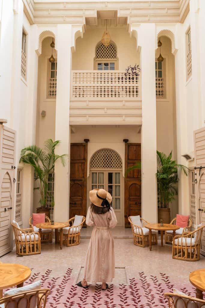 A woman in a pink dress and straw hat stands at the center of a Marrakech riad courtyard, framed by tall white horseshoe arches, carved wooden doors, rattan armchairs, potted palms, and a patterned pink rug.
