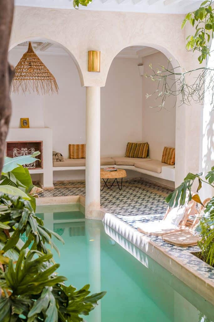 Marrakech riad pool nook with a small green-tiled plunge pool, cream tadelakt walls and arches, built-in benches with mustard cushions, a rattan pendant light, and patterned floor tiles dappled with sunlight.