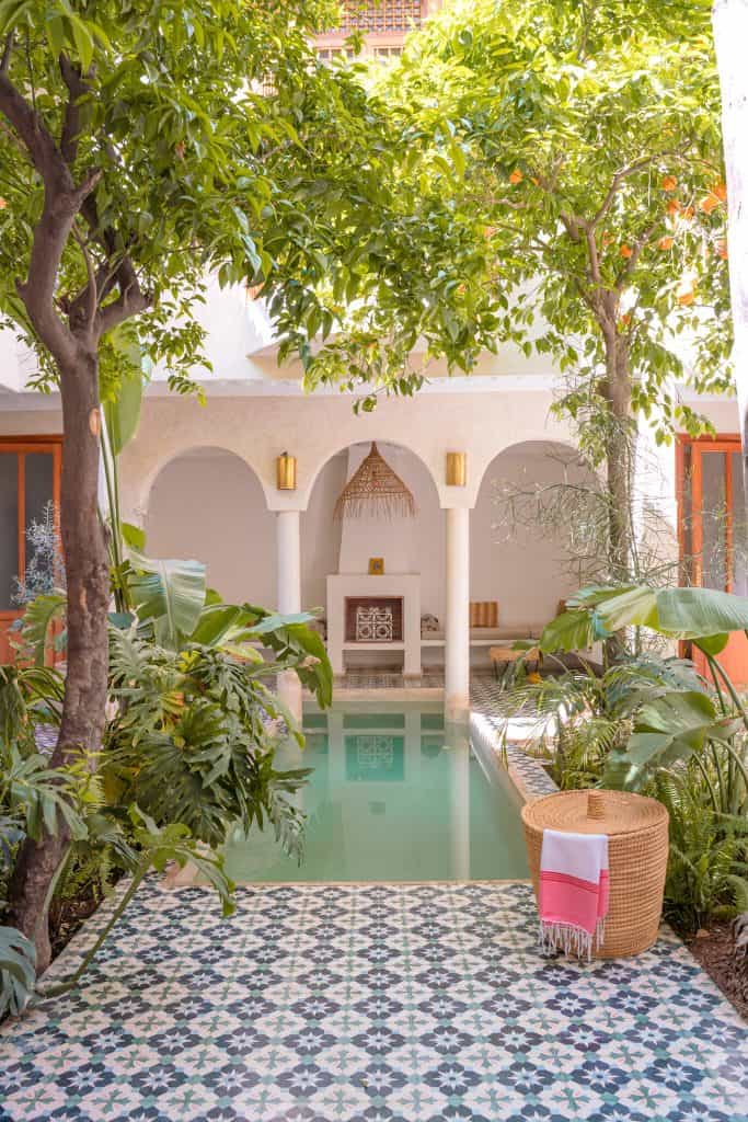 Marrakech riad courtyard with a green-tiled plunge pool flanked by tall citrus trees, white horseshoe arches over a tadelakt seating nook, patterned blue-and-white floor tiles, and a woven basket holding a striped towel.