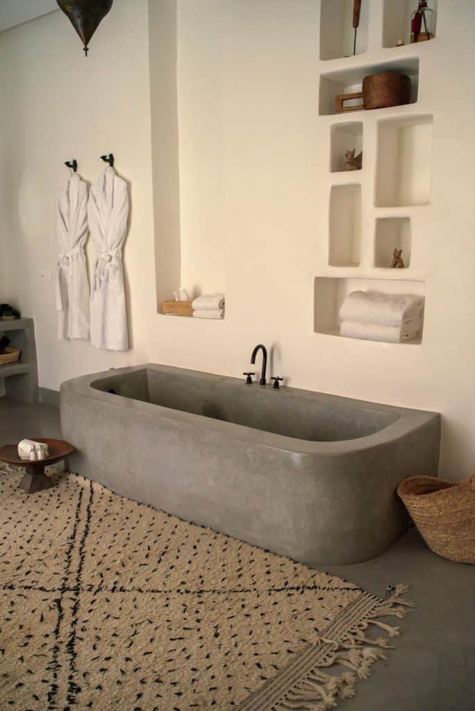 Minimalist Marrakech bathroom with a sculptural concrete soaking tub, white waffle robes hanging on the wall, recessed plaster shelving holding folded towels and small objects, and a black-and-cream Beni Ourain rug on the polished concrete floor.