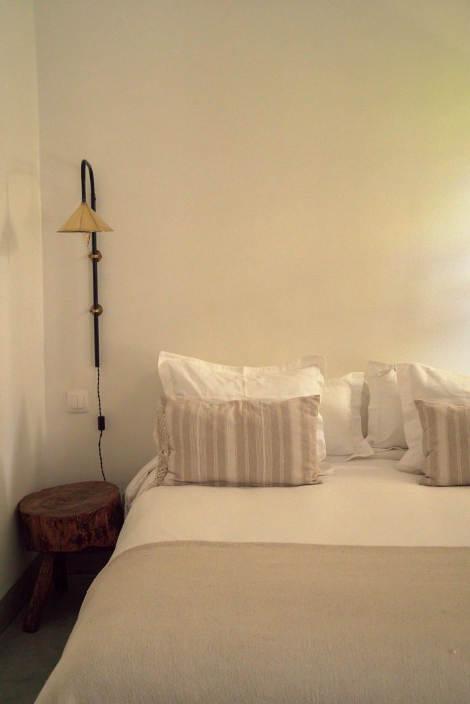 Simple Marrakech guest room bedside detail with a cream wall, a vintage brass wall sconce on a black iron arm, a rustic wooden log stool, and a bed dressed in white linens with striped beige pillows.